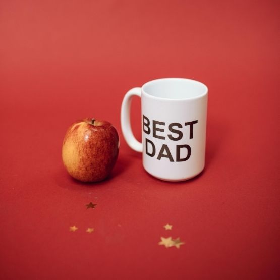 Tasse blanche avec l’inscription "BEST DAD" posée à côté d’une pomme rouge sur fond rouge.