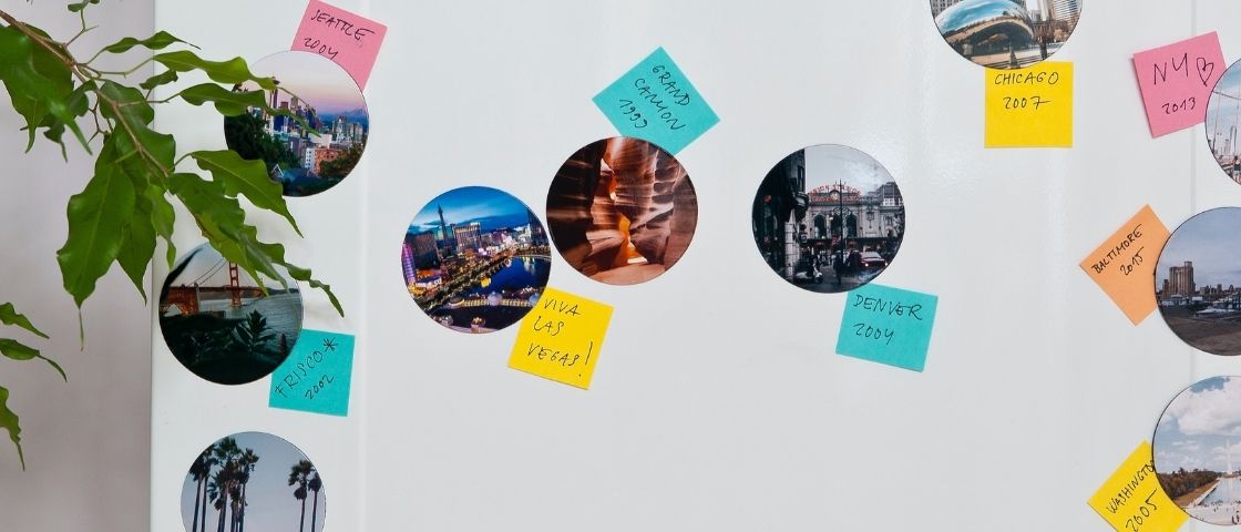 Aimants photo ronds collés sur un frigo avec des post-its colorés indiquant des destinations.