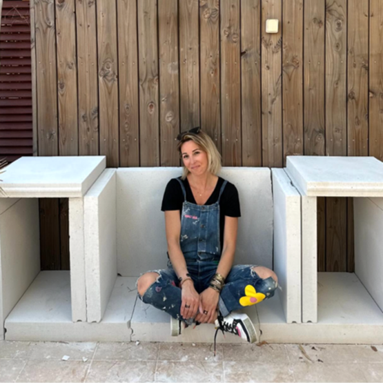 Femme assise sur une structure en béton, en tenue de bricolage, devant un mur en bois.