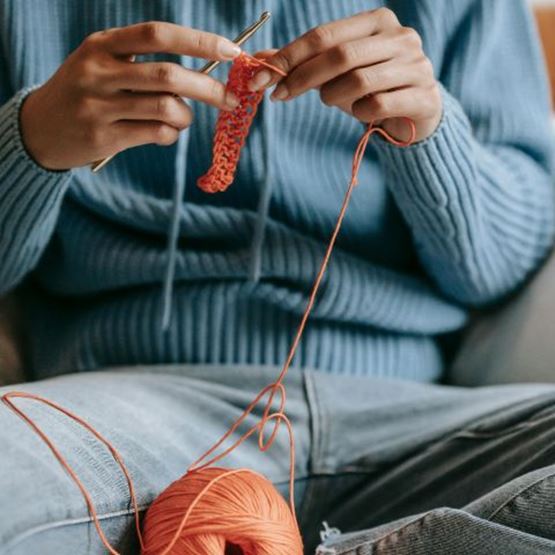 Personne en pull bleu assise, en train de crocheter avec du fil orange.