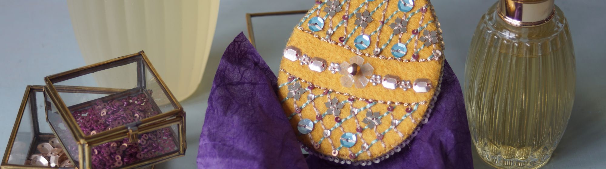 Œuf décoratif en tissu jaune brodé de perles et sequins, posé près de flacons de parfum élégants.