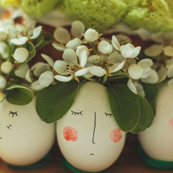 Œufs peints avec des visages et décorés de feuilles vertes et fleurs blanches comme coiffures.