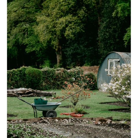 Jardin verdoyant avec une brouette remplie d’outils, devant une cabane en métal arrondie.