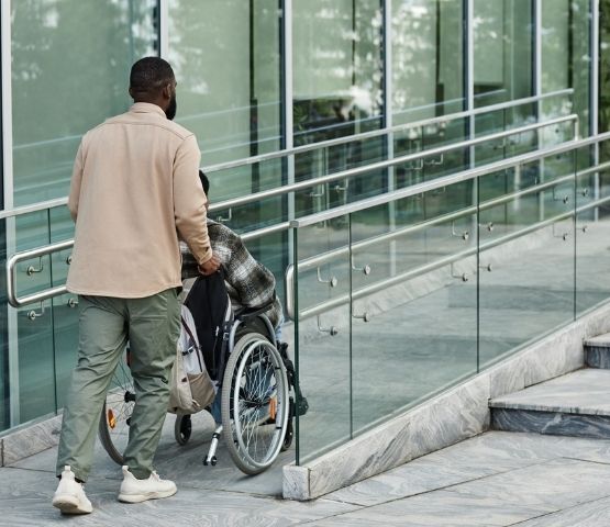 Une personne aide une autre à accéder à un bâtiment en fauteuil roulant, utilisant une rampe d’accès.