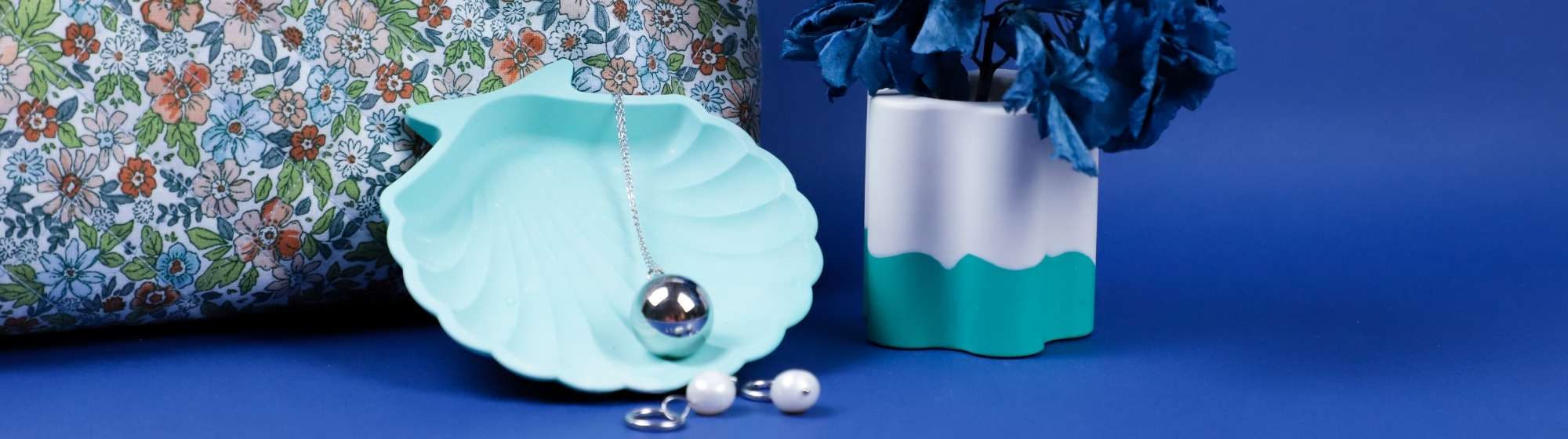 Mise en scène déco avec un pot Jesmonite, un plateau coquillage bleu, des bijoux et un sac à motif fleuri.