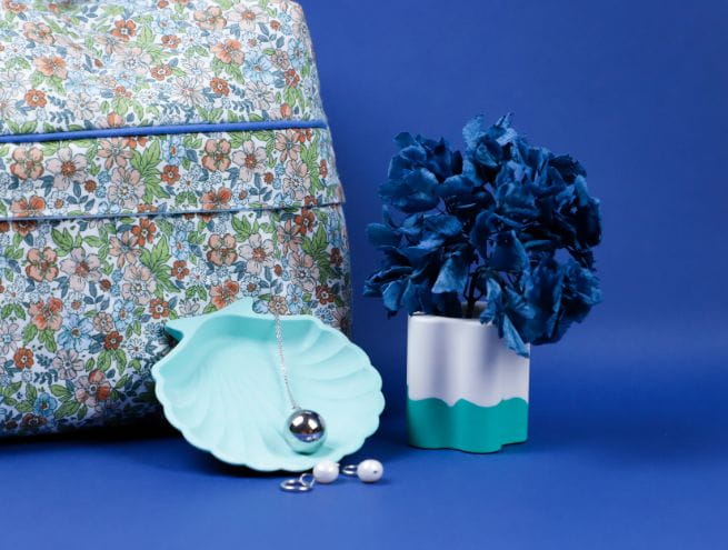Mise en scène déco avec un pot Jesmonite, un plateau coquillage bleu, des bijoux et un sac à motif fleuri.